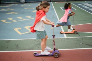 Deux jeunes filles font du scooter de manière ludique sur une aire de jeux colorée, vêtues de capes rouge vif.